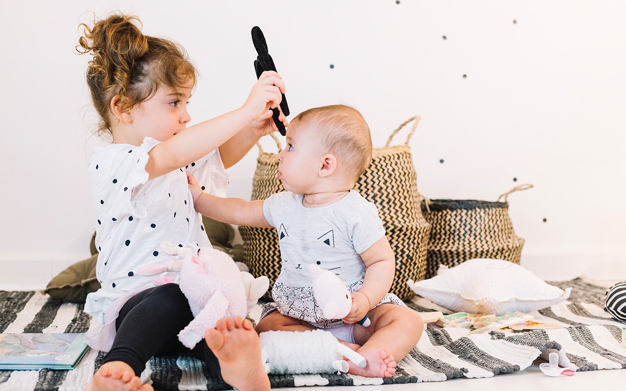 A toddler girl holding a toy up to a baby boy while sitting on a striped rug in a cozy room with woven baskets in the background. Both children are engaged in playful interaction.