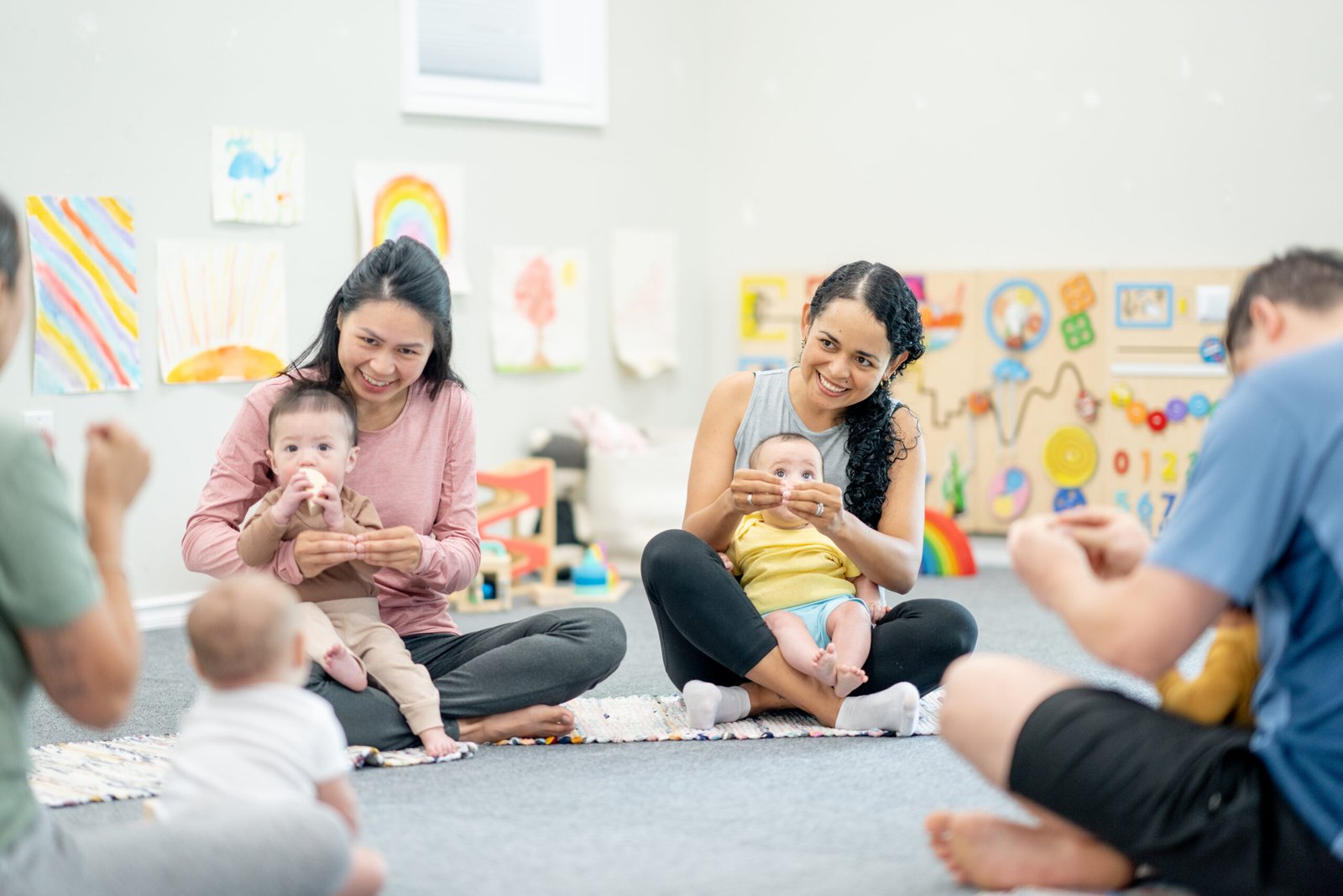 A group of parents and babies sitting on the floor in a bright and colorful classroom, engaging in a baby sign language class. The mothers are smiling while demonstrating hand signs to their infants, who are attentively watching. The background features children's artwork, toys, and educational decorations.