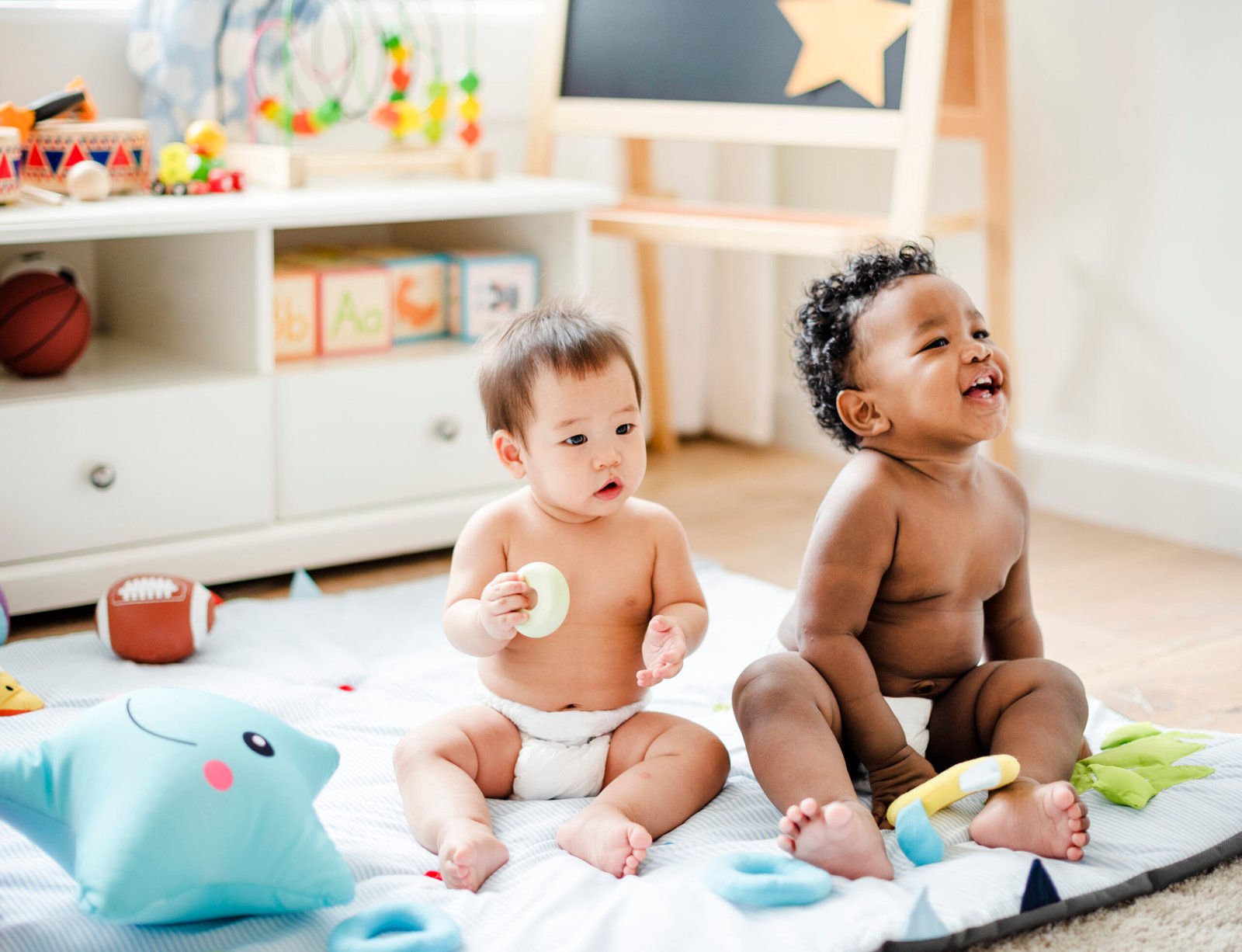 Two babies sitting on a soft play mat, one happily laughing while the other explores a teething toy, surrounded by colorful sensory toys in a bright and inviting playroom.