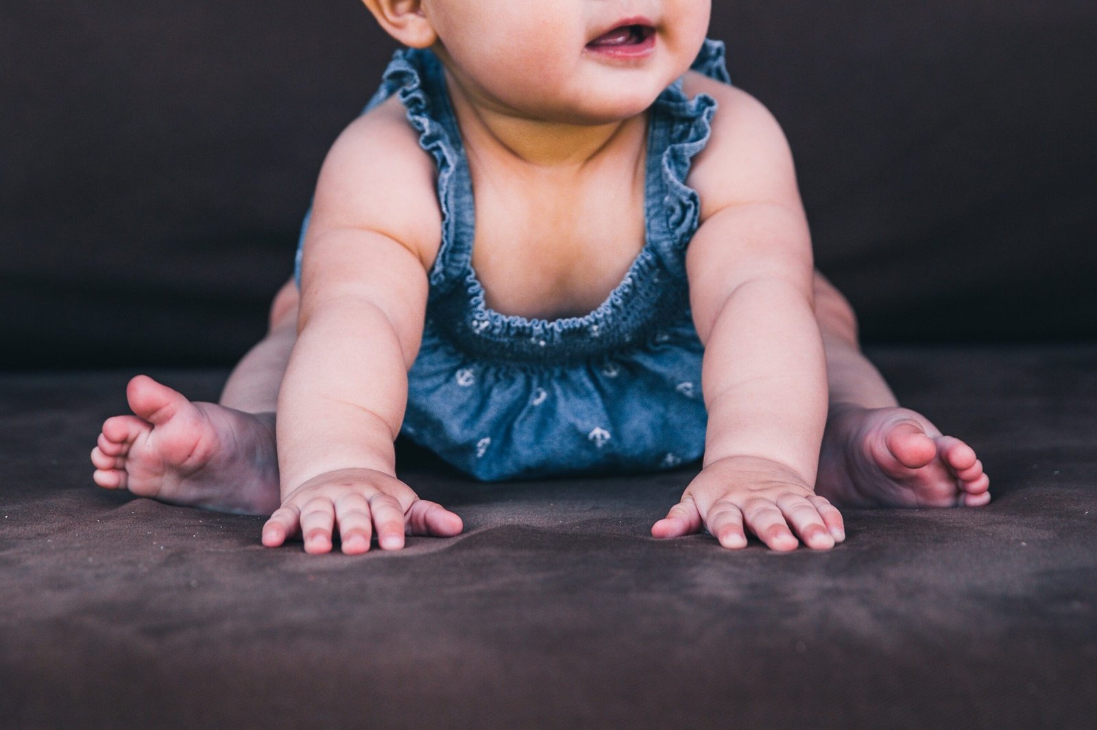 A close-up of a baby in a denim romper practicing tummy time, with arms stretched forward and tiny fingers pressing into a soft surface, building strength and coordination.