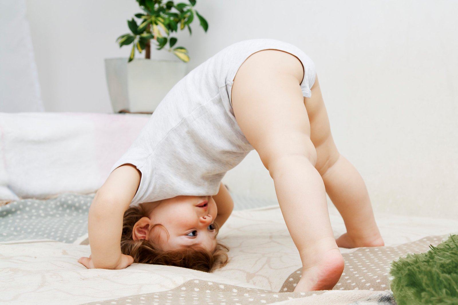 A playful baby in a onesie bends over, curiously peeking through their legs while exploring movement and balance on a soft, surface.