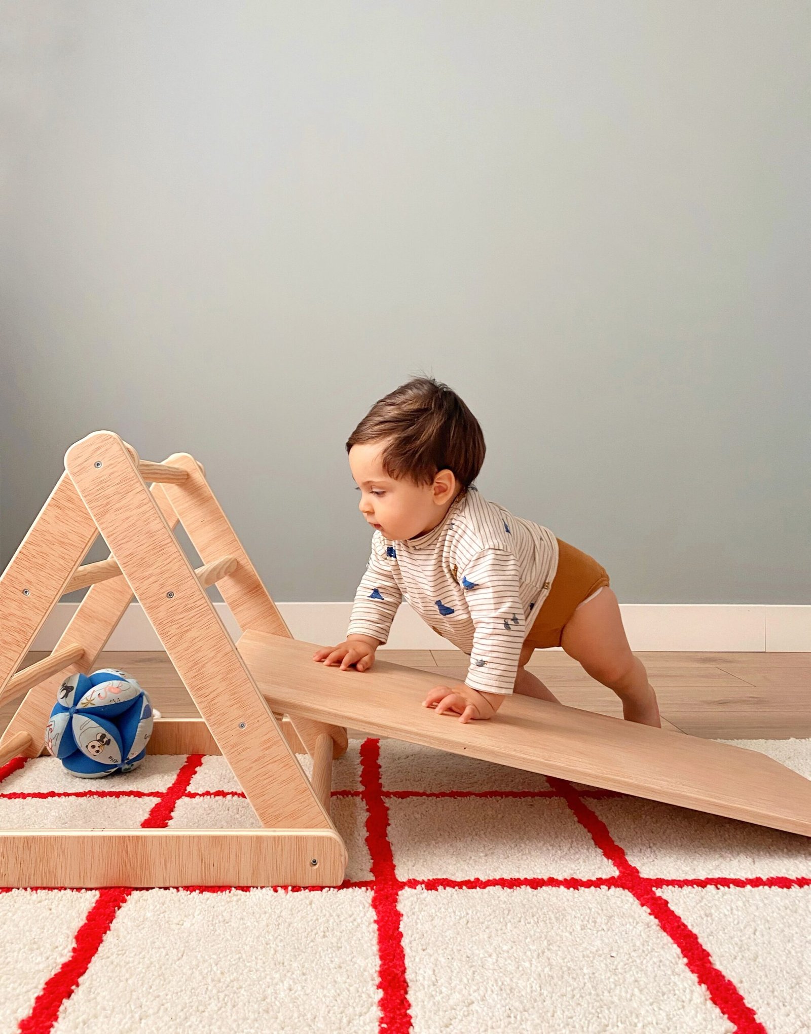 A determined baby boy climbs a wooden Pikler triangle with a ramp, building strength, coordination, and confidence through active play. The setup encourages gross motor development and independent exploration.