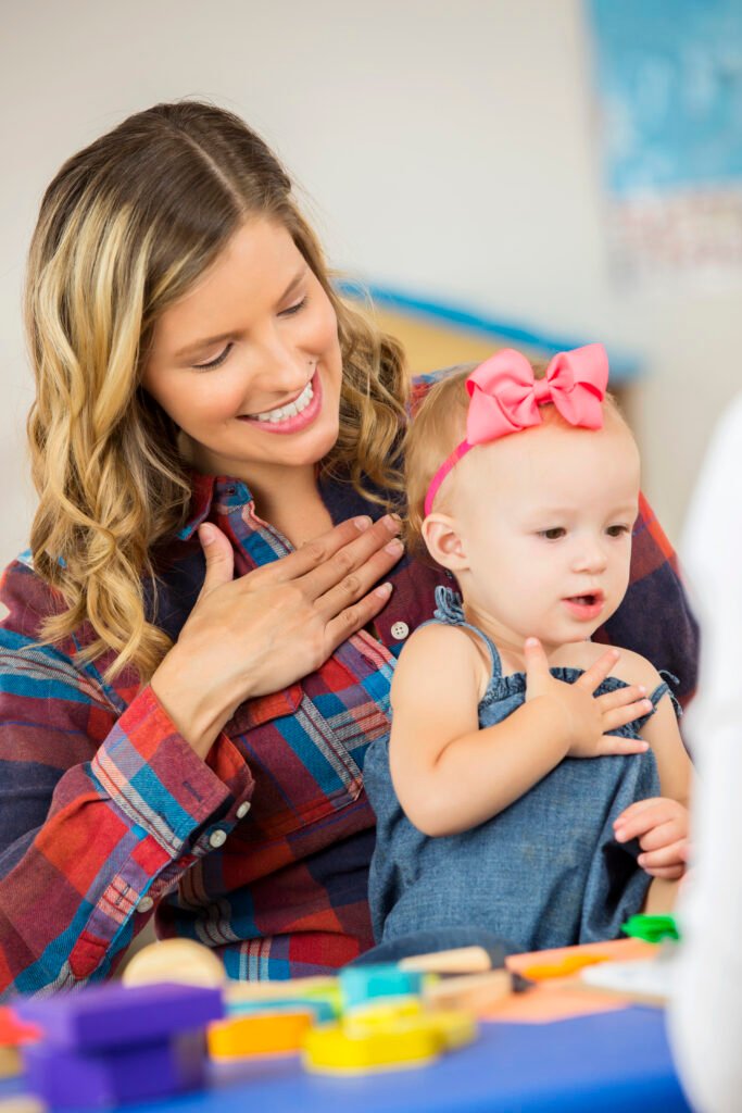 A mother and her baby girl are engaging in a baby sign language activity. The mother, wearing a plaid shirt, smiles warmly as she places her hand on her chest, modeling the sign for "please." The baby girl, dressed in a denim outfit with a bright pink bow, mimics her mother's gesture with a focused expression. Colorful toys and learning materials are visible in the foreground, suggesting an interactive learning environment.
