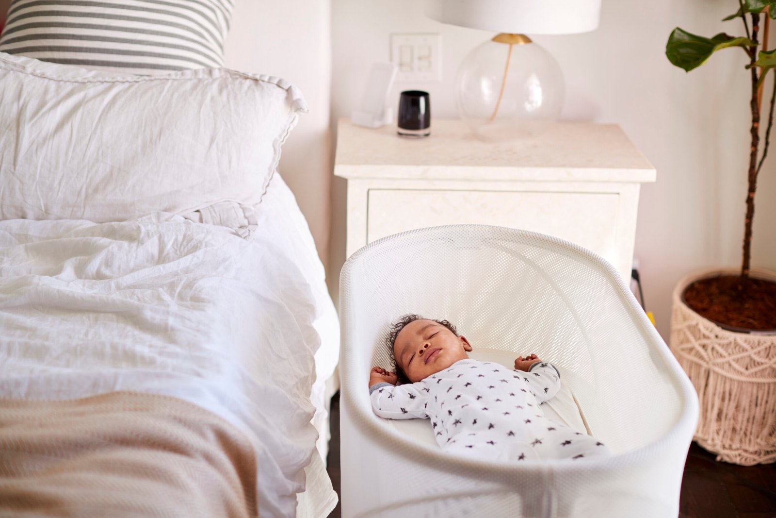 A newborn baby sleeps peacefully in a white bassinet next to a cozy, neatly made bed with soft linens and pillows. The baby is dressed in a white onesie with tiny patterns, resting with arms slightly raised. A nightstand with a lamp and a small plant in a woven basket are visible in the background, creating a warm and serene bedroom environment.
