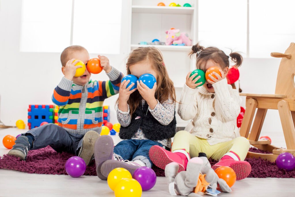 Three young children sitting on a purple rug in a bright playroom, holding colorful plastic balls over their eyes while laughing and playing together. The room is filled with toys, including a rocking horse, building blocks, and scattered play balls, creating a fun and engaging environment.