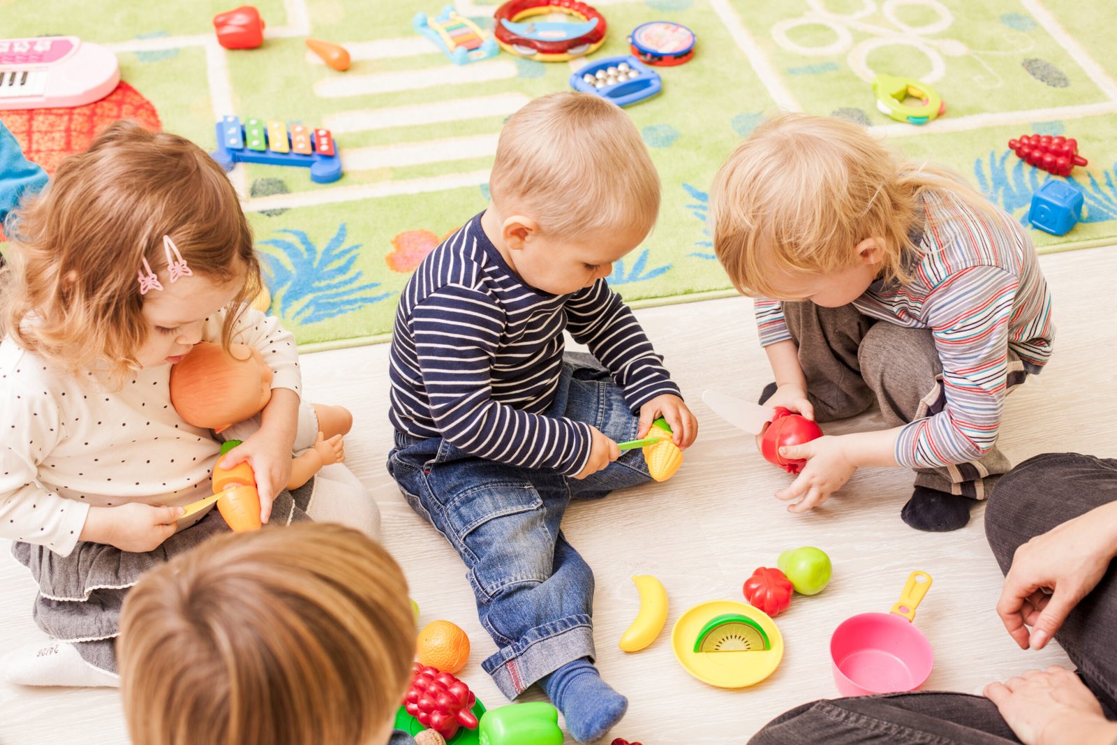 A group of young children sitting on the floor engaged in pretend play with toy food items, cutting plastic fruits and vegetables while role-playing in a play kitchen environment.