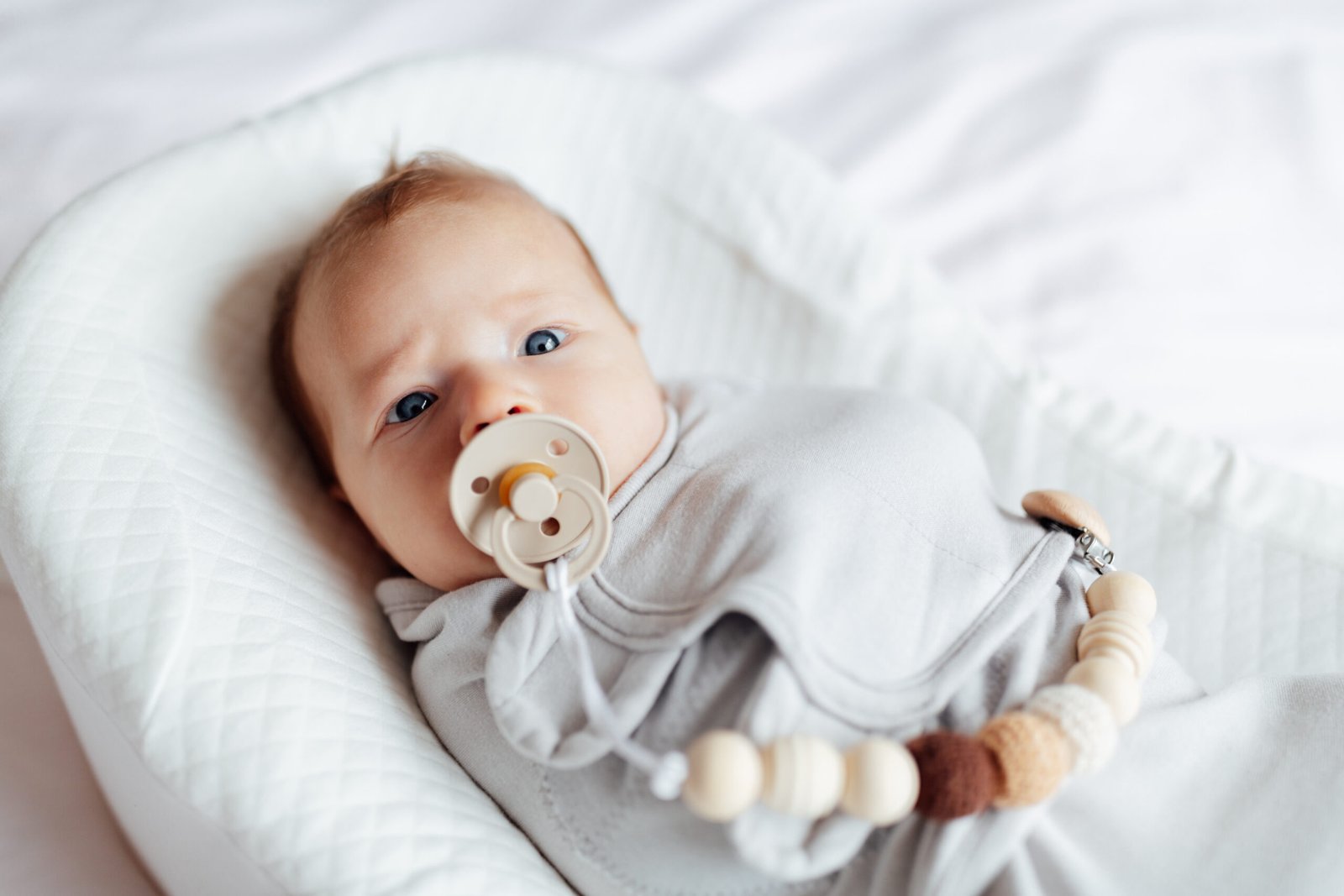 A newborn baby swaddled in a soft gray blanket lies comfortably in a cushioned baby lounger. The baby has wide, curious blue eyes and is sucking on a beige pacifier attached to a beaded clip. The neutral color palette and gentle lighting create a serene and cozy atmosphere, emphasizing the baby's peaceful and content expression.