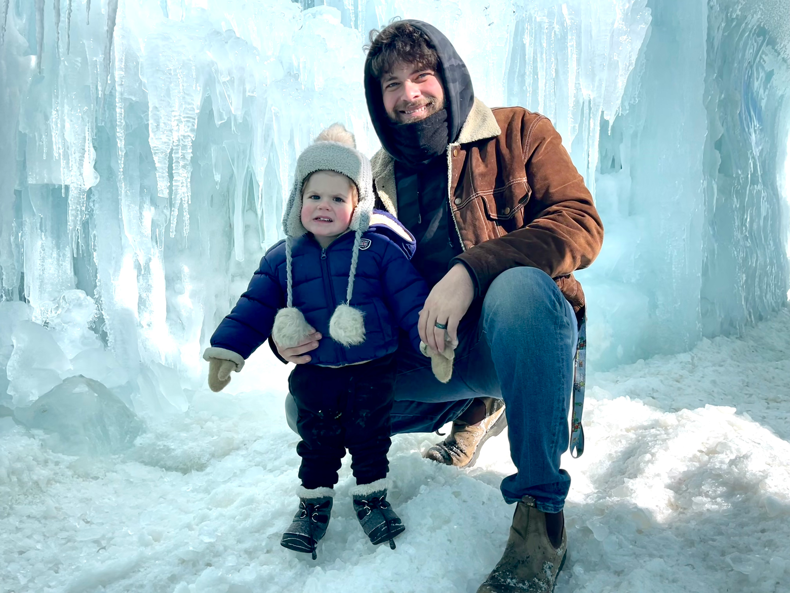 A smiling father with curly brown hair and a beard, dressed in a brown suede jacket, blue jeans, and boots, crouches down next to his toddler son in a winter setting. The little boy, wearing a navy blue puffer jacket, black snow pants, a gray hat with fluffy earflaps, and mittens, looks slightly chilly but happy. They are surrounded by towering ice formations and icicles, with snow covering the ground, creating a magical frozen landscape.