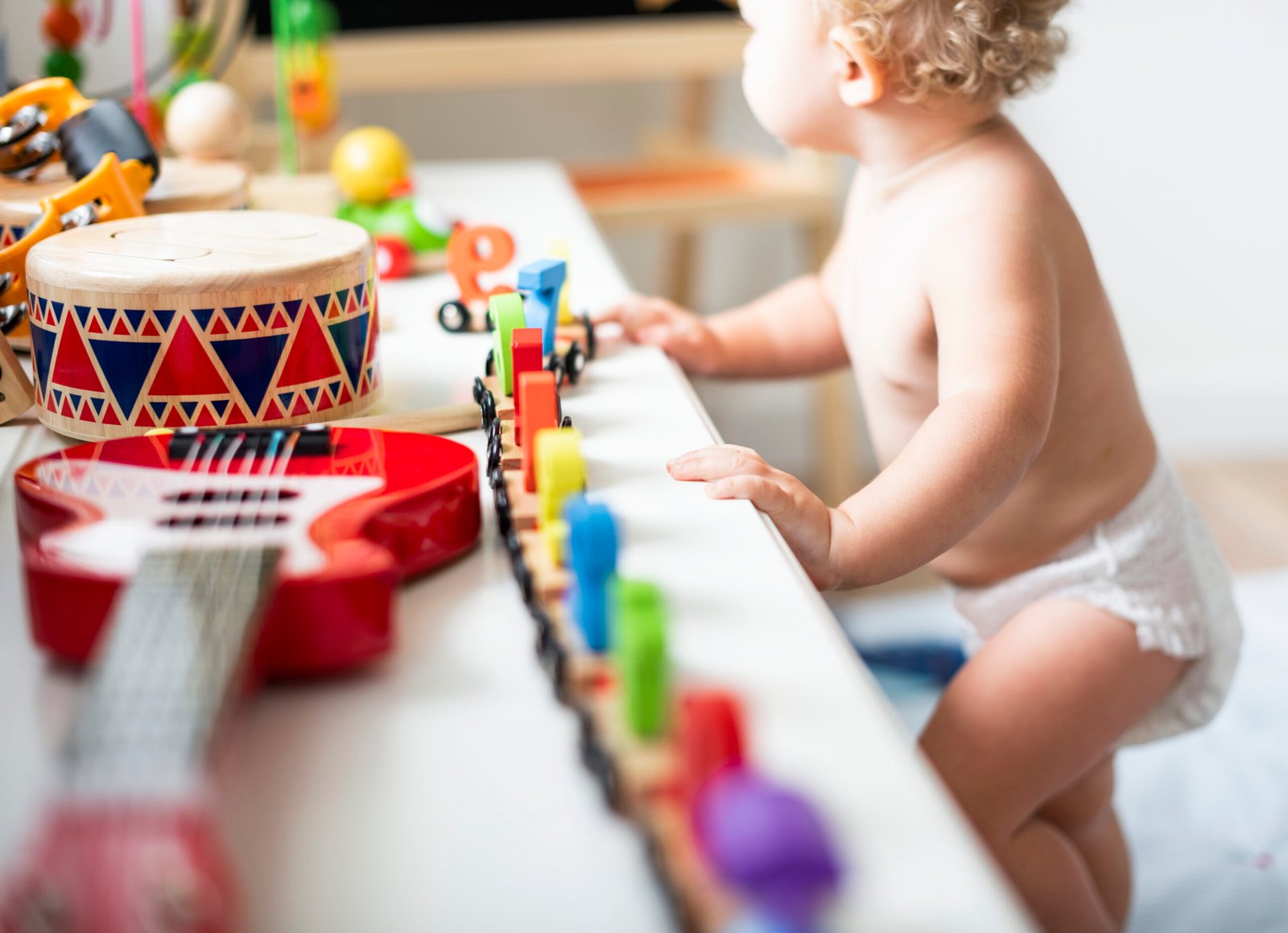A young child in a diaper stands at a play table, reaching out to explore a colorful wooden train set and musical instruments. A red toy guitar and a decorative wooden drum with vibrant geometric patterns are visible in the foreground. The scene is filled with sensory-rich toys, encouraging creative and musical play in a bright, engaging environment.