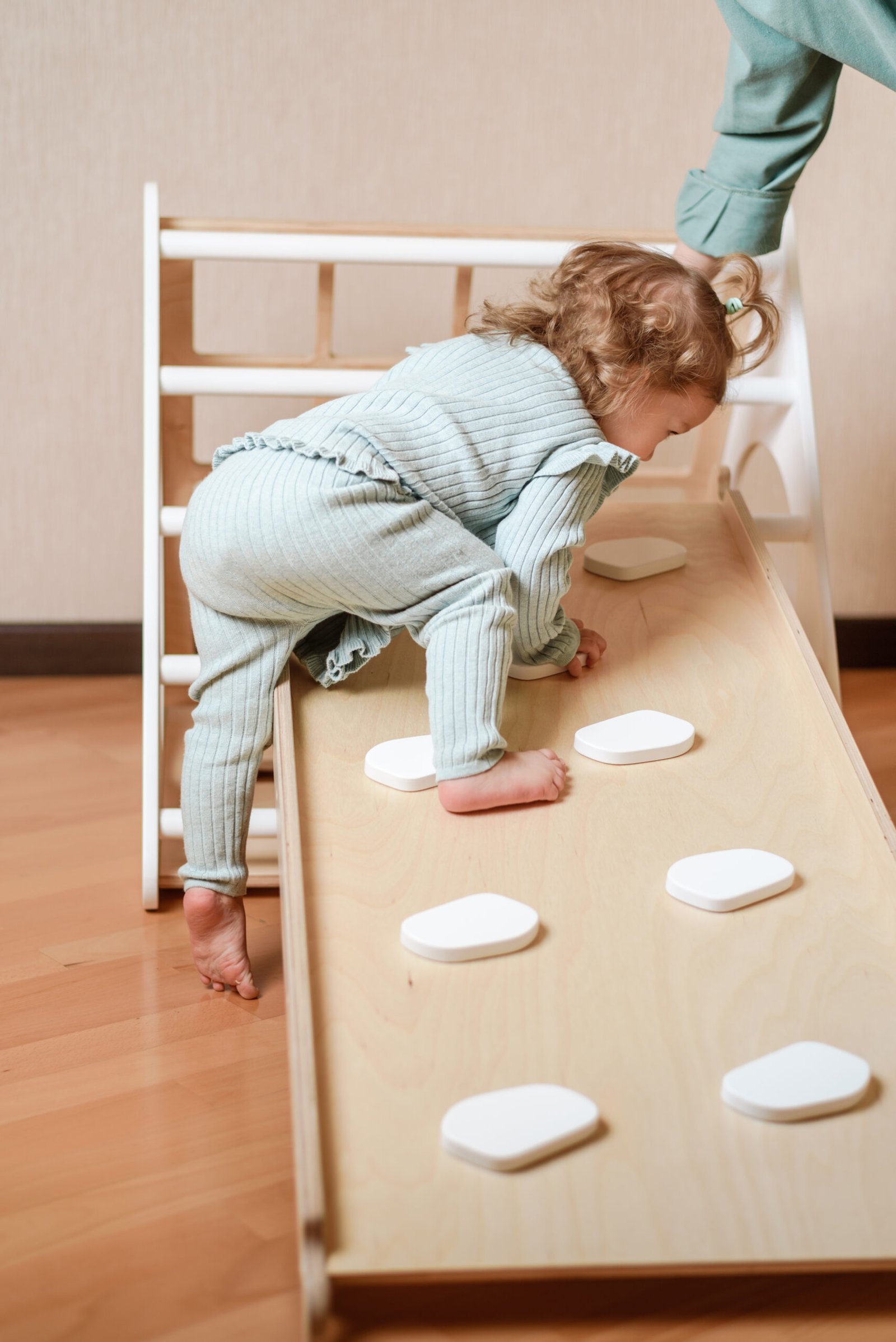 A young toddler girl in a ribbed light green outfit climbs a wooden Montessori-style slide with white stepping stones, carefully making her way up. A caregiver's hand is visible at the top, offering support. The indoor play area has a wooden floor and neutral-colored walls.