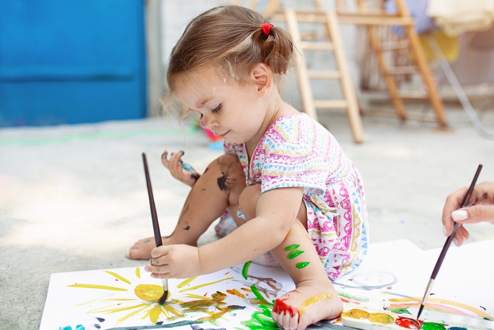 A young girl with pigtails sits on the ground, fully immersed in a painting activity. She holds a paintbrush, adding colorful strokes to her artwork, while her hands, legs, and feet are covered in paint. She wears a patterned dress, and her expression shows deep concentration. An adult’s hand, also holding a paintbrush, can be seen assisting her. The outdoor setting, with an easel and art supplies in the background, enhances the creative atmosphere.