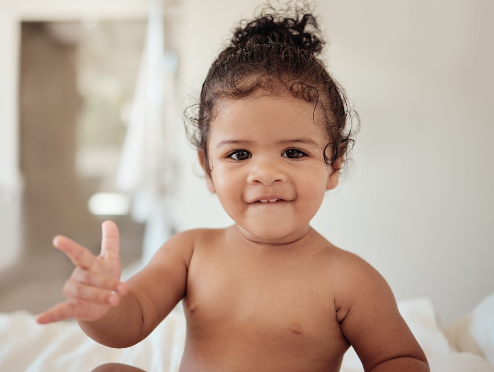 A young girl with curly hair and a bright smile sits on a bed, looking directly at the camera. She holds up her hand, forming the "I love you" sign in American Sign Language. Her expression is warm and playful, with a slight bite of her lower lip, capturing a moment of connection and communication. The soft, natural lighting and blurred background create a cozy and intimate atmosphere.