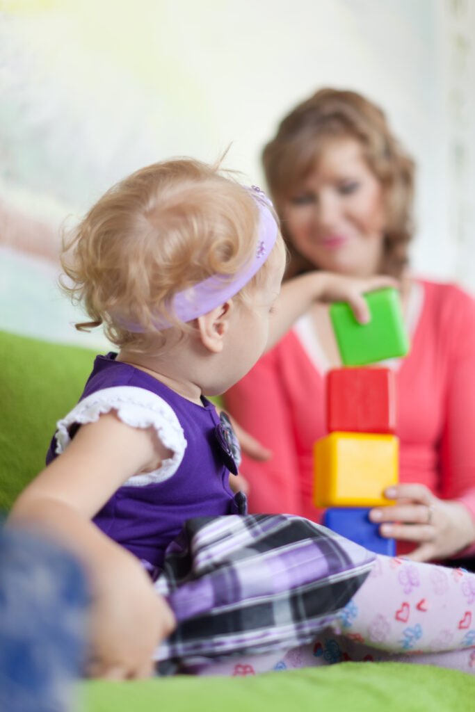 Little girl in purple dress and purple headband stacking blocks while her mother holds the block sturdy