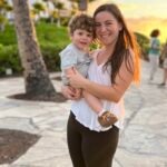A smiling mother with long brown hair, wearing a white tank top, black leggings, and fuzzy slippers, holds her curly-haired toddler boy in her arms. The boy, dressed in a light gray t-shirt and shorts, looks at the camera with a happy expression. The background features a tropical setting with palm trees, green bushes, and people walking along a stone pathway, with the golden glow of the setting sun casting warm light over the scene.