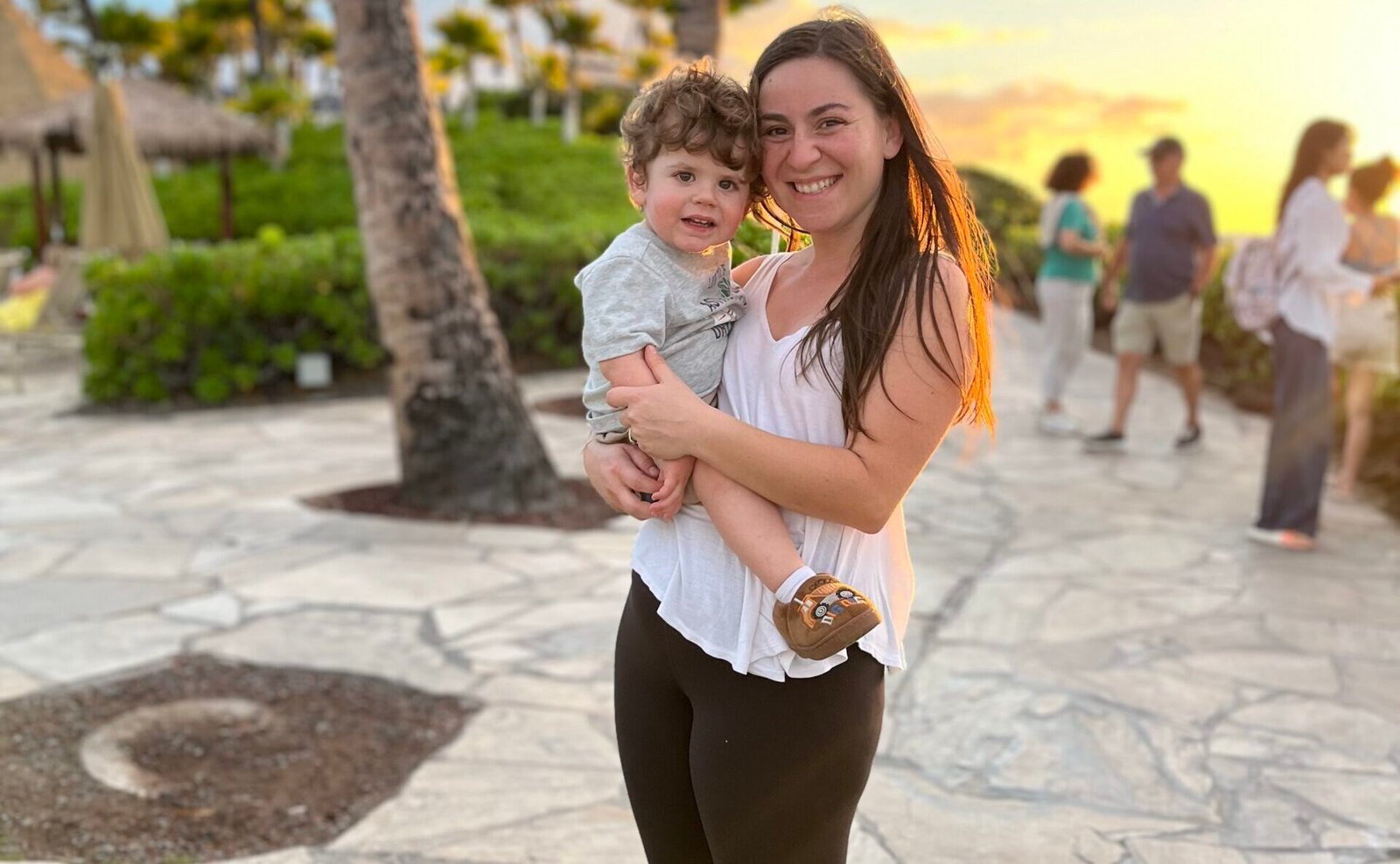 A smiling mother with long brown hair, wearing a white tank top, black leggings, and fuzzy slippers, holds her curly-haired toddler boy in her arms. The boy, dressed in a light gray t-shirt and shorts, looks at the camera with a happy expression. The background features a tropical setting with palm trees, green bushes, and people walking along a stone pathway, with the golden glow of the setting sun casting warm light over the scene.