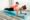 A mother in athletic wear doing a forearm plank on a blue yoga mat while her young son playfully lies on her back. Both are smiling, enjoying an active bonding moment in a bright home setting.