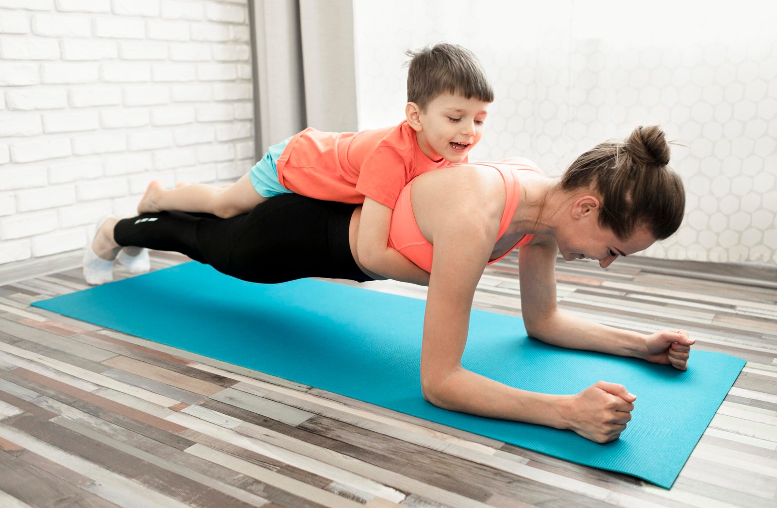 A mother in athletic wear doing a forearm plank on a blue yoga mat while her young son playfully lies on her back. Both are smiling, enjoying an active bonding moment in a bright home setting.