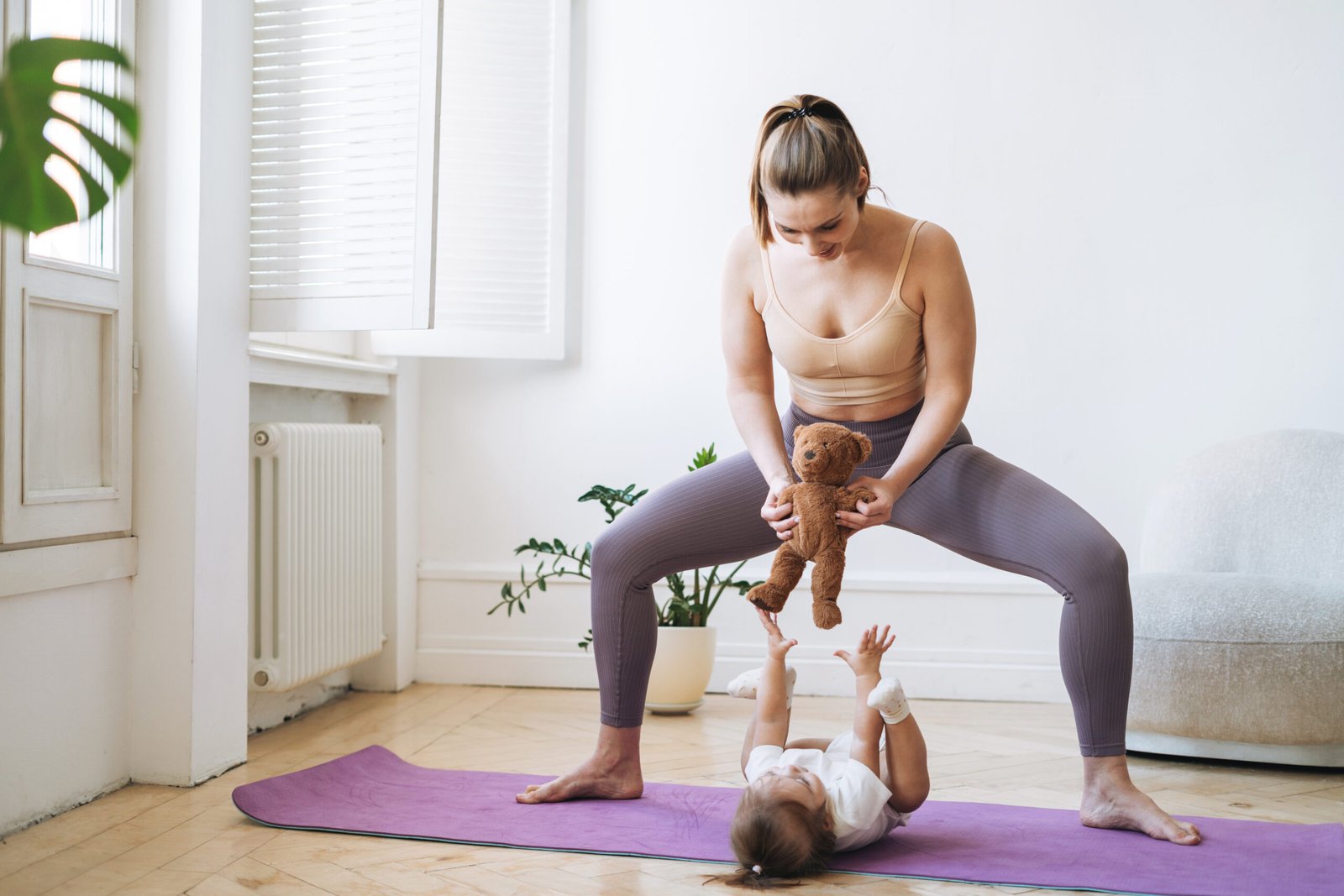 A mother in workout clothes performing a sumo squat on a purple yoga mat while holding a teddy bear above her baby, who is lying on the mat reaching up with excitement. The scene is set in a bright and airy living space.