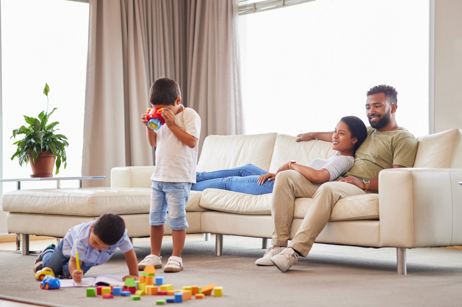 A smiling couple sits together on a cream-colored couch in a bright living room while their two young children play nearby. One child is kneeling on the carpet, drawing and playing with colorful building blocks, while the other stands holding a toy camera. The scene conveys a warm and relaxed family atmosphere.