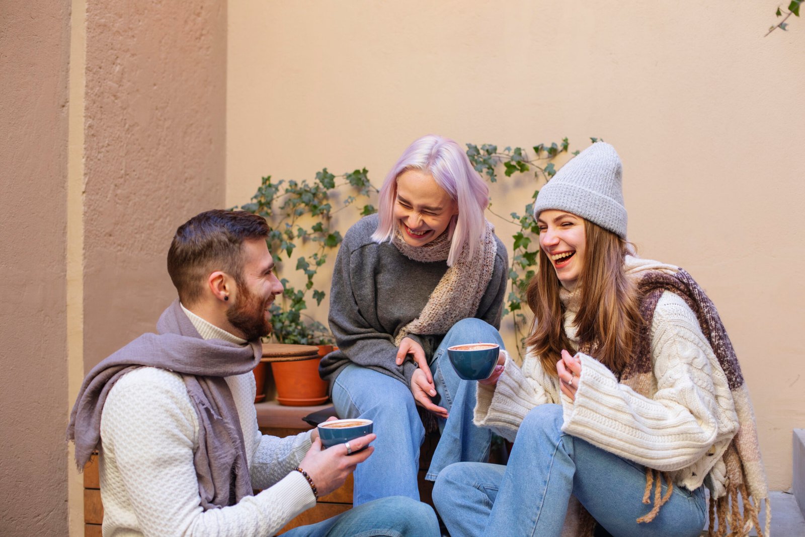 A group of three friends sitting outdoors, dressed in cozy winter clothing, laughing and enjoying warm drinks together.