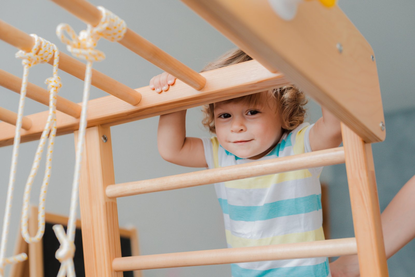 A young toddler boy with curly hair and a striped shirt climbs a wooden jungle gym, gripping the bars while smiling confidently. The indoor play structure features ropes and wooden beams, encouraging active play.