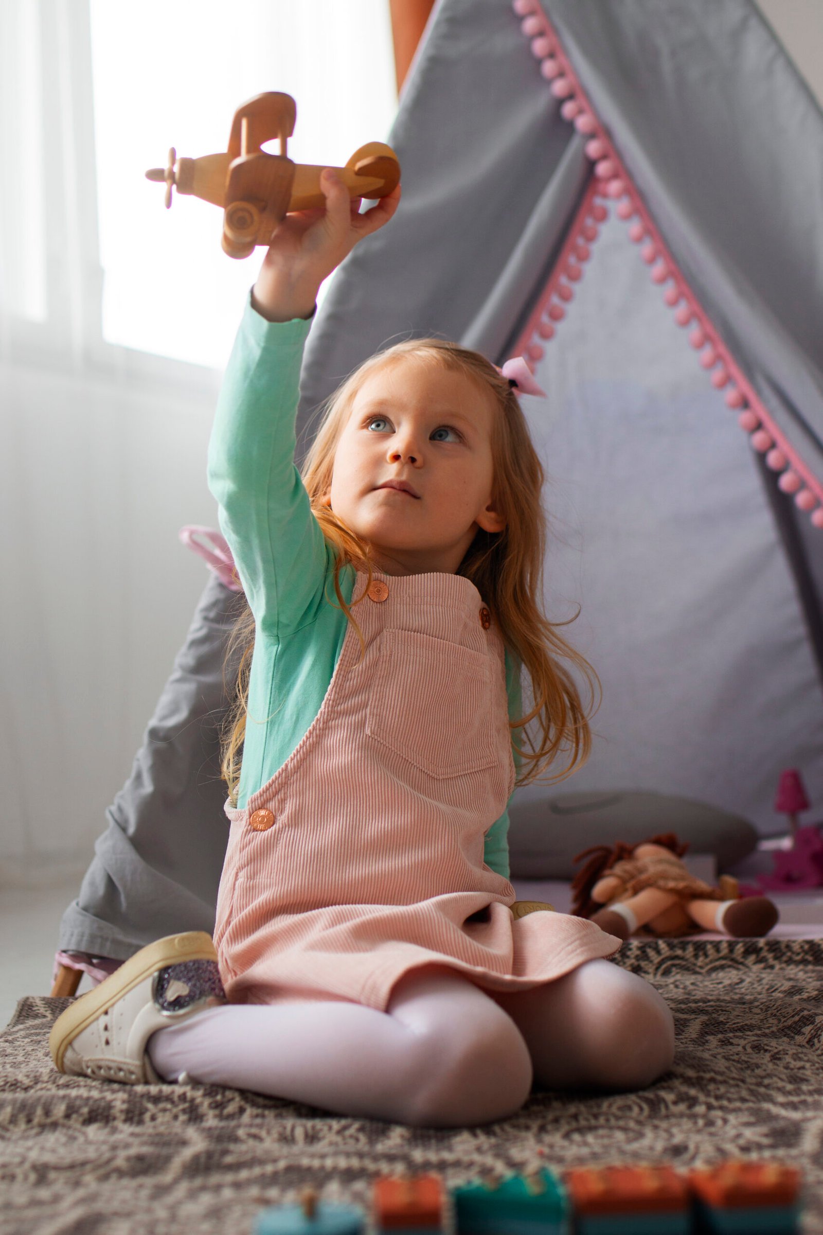 A young girl with long blonde hair sits on a rug, holding a wooden toy airplane high in the air while gazing up at it with wonder. She is dressed in a pink corduroy pinafore, a mint-green long-sleeve shirt, and white tights. Behind her, a cozy play tent decorated with pink pom-pom trim creates a whimsical play space filled with stuffed animals and toys.