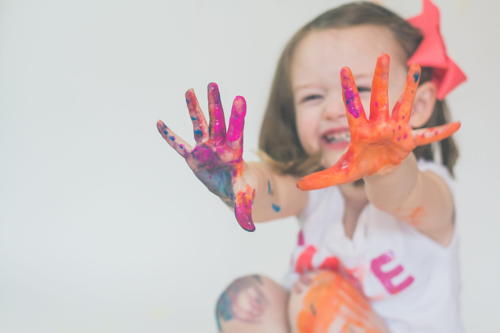 A joyful toddler with a bright pink bow proudly showing her colorful, paint-covered hands, fully immersed in a fun and creative art activity.