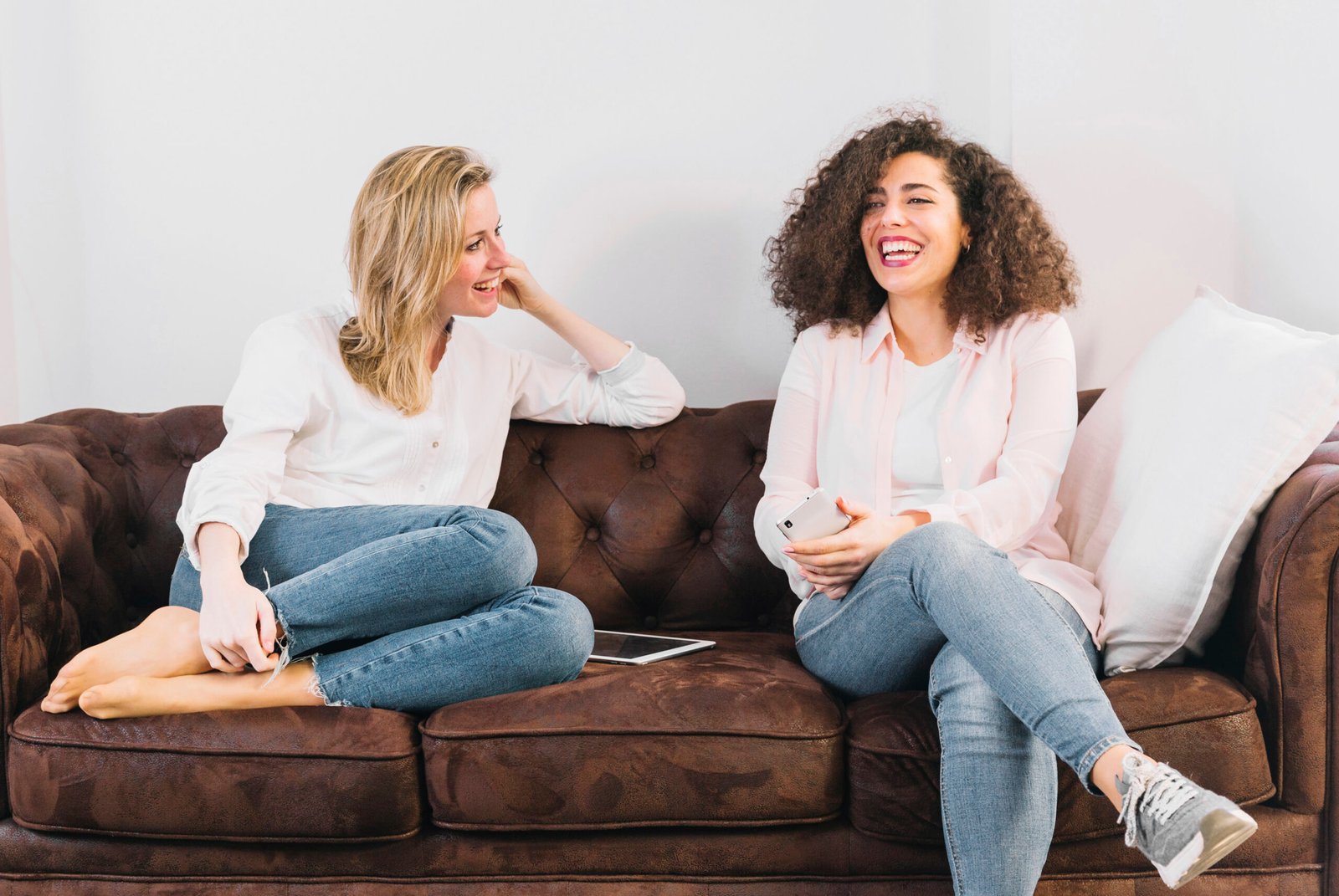 Two women sitting on a brown couch, laughing and enjoying a conversation, creating a warm and friendly atmosphere.