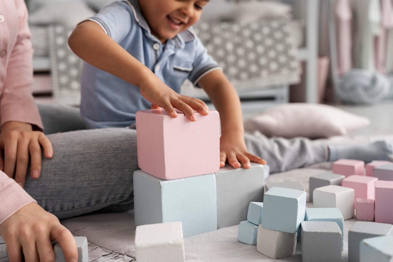 A young child excitedly stacks pastel-colored wooden blocks while sitting on the floor, engaging in hands-on play. Their face lights up with joy as they carefully balance the pieces, developing fine motor skills, hand-eye coordination, and problem-solving abilities.