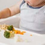 Baby sitting in his highchair, eating broccoli, cauliflower and squash by himself