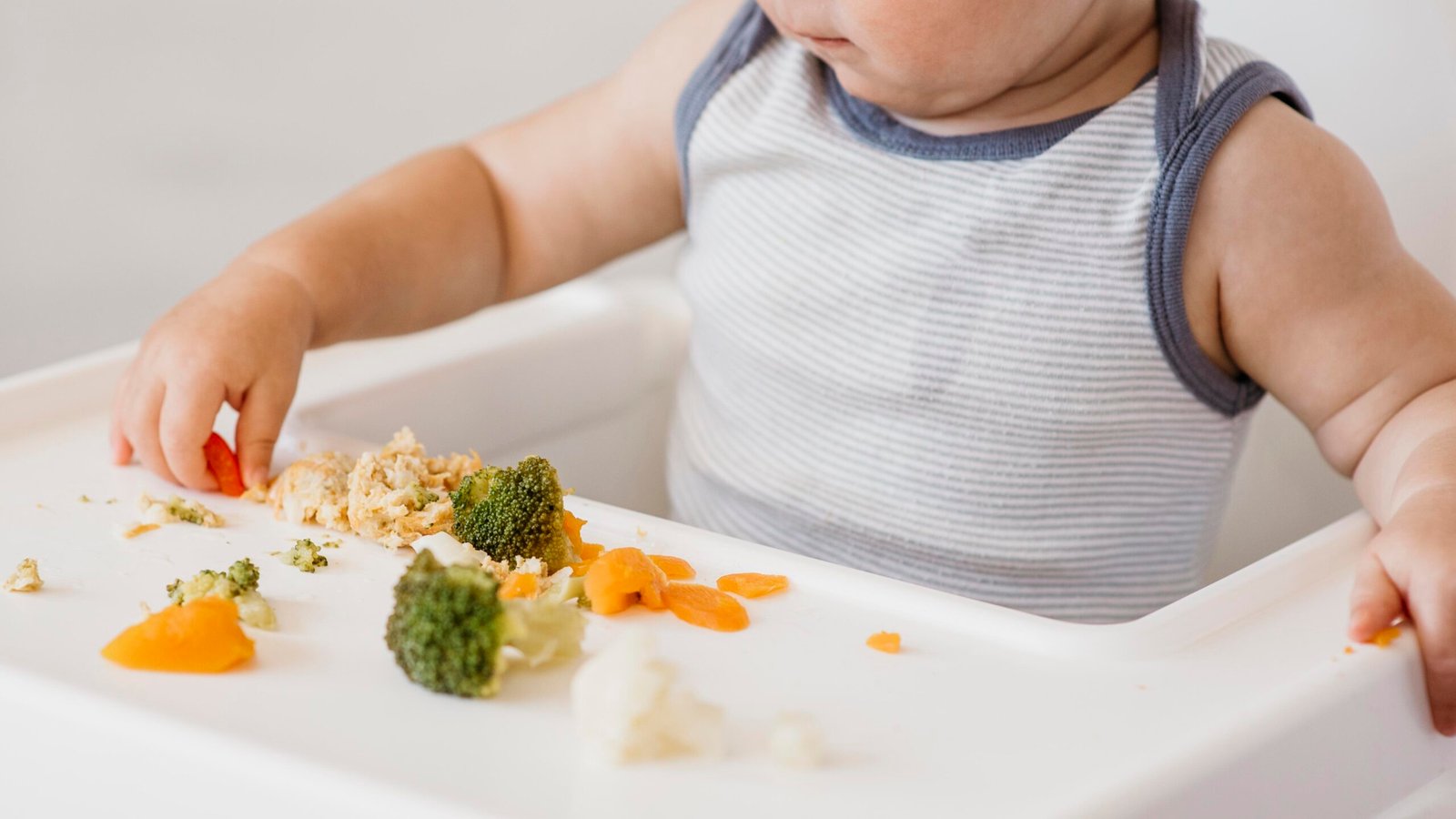 Baby sitting in his highchair, eating broccoli, cauliflower and squash by himself