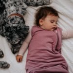 Baby girl sleeping next to a dog wearing a pink sleep sack.