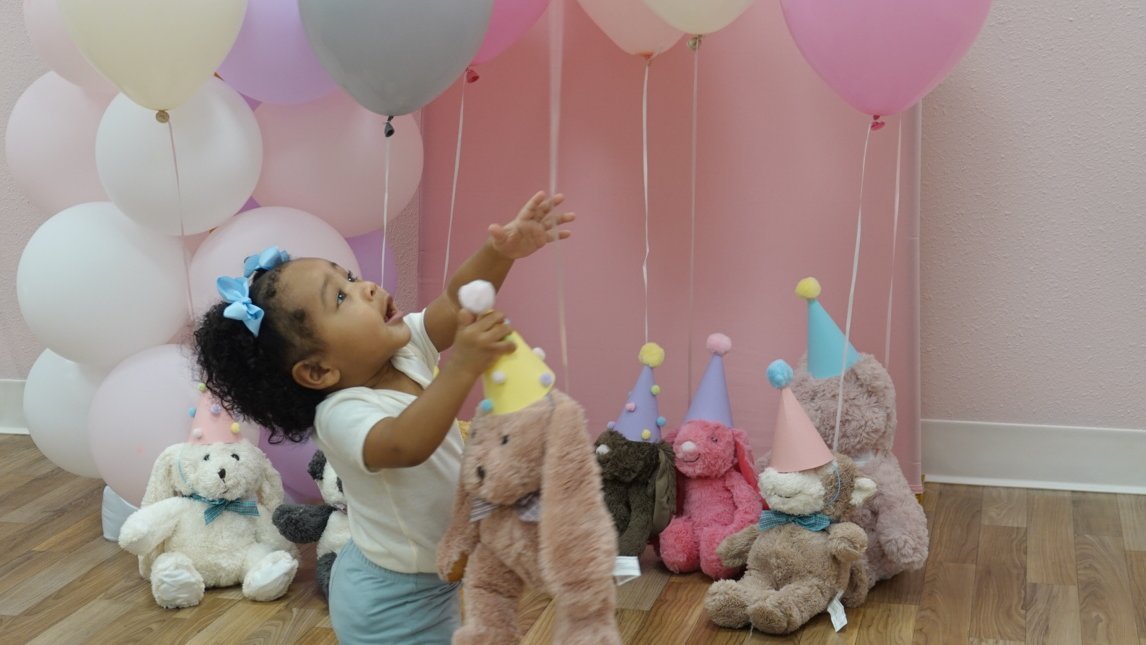 A toddler reaching for balloons surrounded by party-hat stuffed animals at a Baby University birthday party