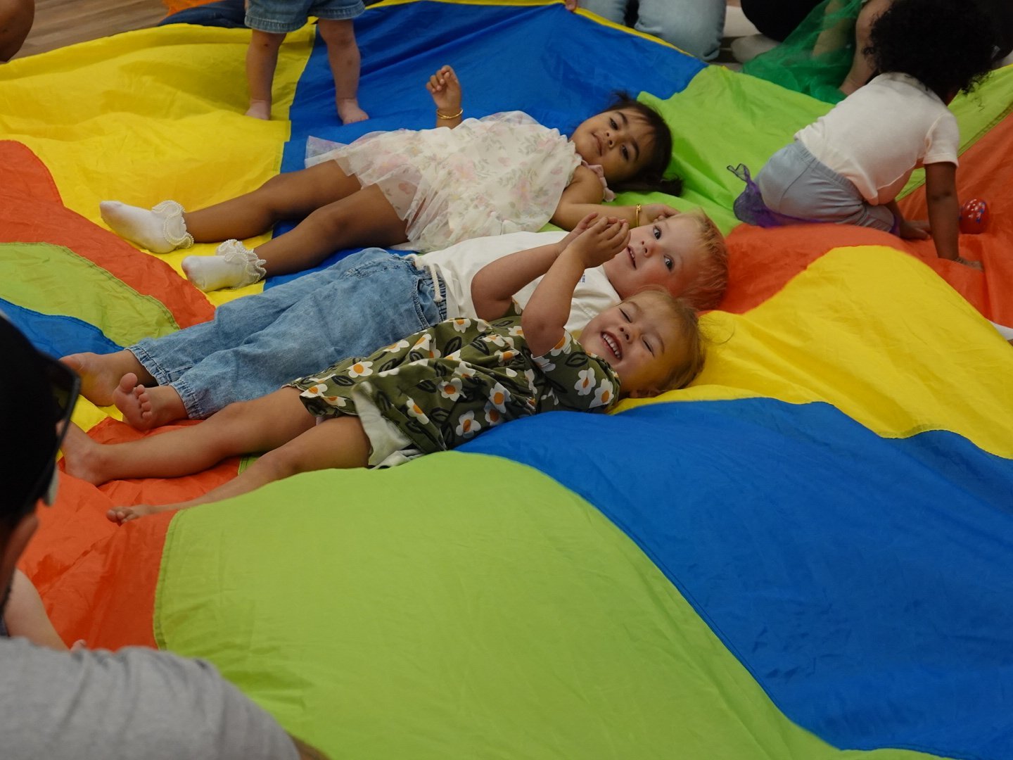 Toddlers laughing on a rainbow parachute during music class at Baby University