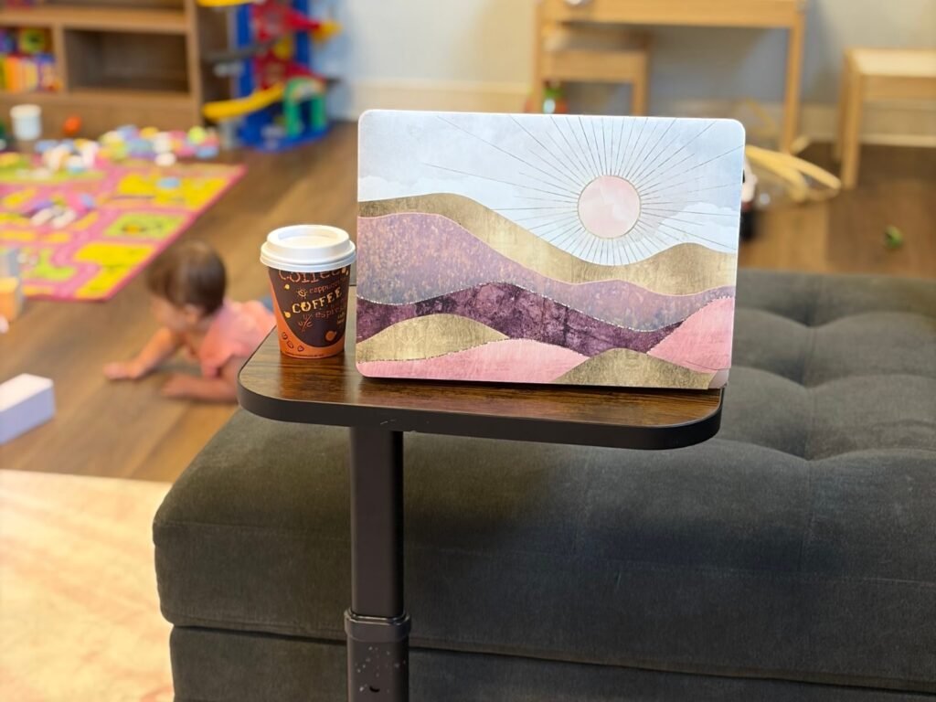 Laptop and coffee on a side table by the lounge couch while a baby plays nearby at Baby University